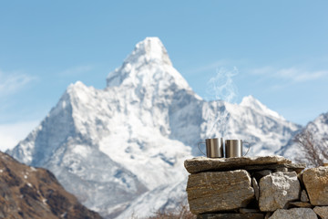 Two cups are on the stones in focus. Steam from cups. Mount Ama Dablam is blurred. Everest trekking...