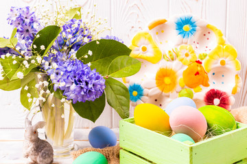 Colorful Easter eggs in a green box and rabbit figurine and spring bouquet in a vase and a beautiful ceramic plate on a white wooden table