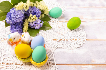 Colorful Easter eggs in a ceramic rabbit stand and a spring bouquet with lilac and yellow flowers on a beautiful knitted napkin on a white wooden table