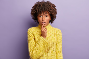 Confused focusd young woman keeps hand near mouth, expresses wonder as listens something surprising, wears yellow jumper, has dark healthy skin, Afro hairstyle, isolated on purple background