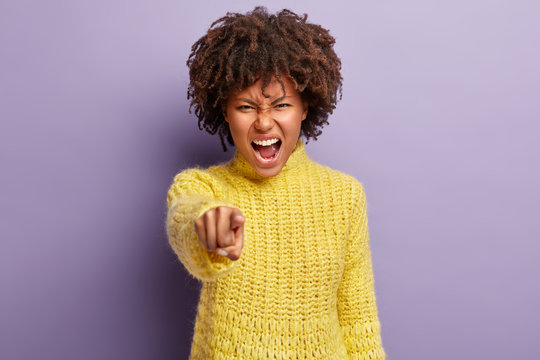 Annoyed pretty woman with curly hair accuses you, points straight at camera, being mad, blames her failures, shouts loudly from anger, isolated over purple background. You are guilty in my fault