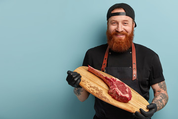 Studio shot of cheerful bearded young man holds wooden board with piece of raw meat, wears black rubber gloves, going to cook supper in restaurant, works in food industry sphere. Cooking concept
