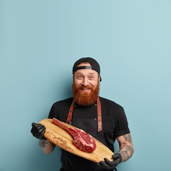 Vertical shot of glad bearded young man with thick ginger beard, holds cutting board with fresh...