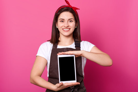 Photo of young attractive female stands with toothy smile and shows new gadget isolated over pink background. Model dresses t shirt, apron, hair band, poses, demonstrates blank screen on mobile phone.