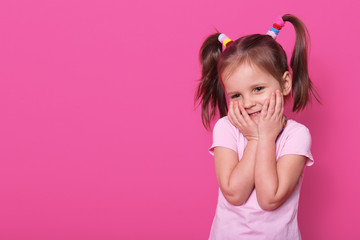 Charming positive small child with two pony tails and many colourful scrunchies, dressed in rose t...