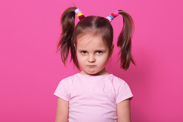 Sad little girl stands against pink wall, looks at camera. Cute kid wears rose t shirt, has two fanny poni tails with many colourful scrunchies, looks hurt with pouty lips. Upset child on playground.