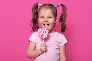 Happy little girl with striped lollypop in shape of heart in hand. Charming kid wears casual rose t...