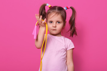 Close up portrait of cute, sad girl holds in hand skipping rope. Little child wants to play with somebody. Adorable kid with pony tails and colourful scrunchies, wears t shirt against rose background.