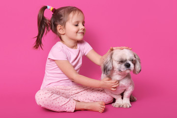 Little girl pets her pekingese while sitting with crossed legs on floor. Adorable child likes her...
