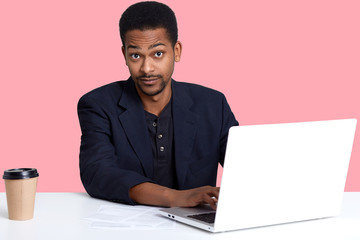 Indoor portrait of young handsome black man sitting at table, drinks coffee or tea and works with lap top. Model looks surprised at camera while typing on keyboard, expreses astonishment with face.