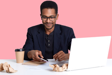 Handsome African American male sits at desk with lap top and crumpled shirts of paper, works online. Dark skinned guy takes smart phone and looks down on it, drinks coffee or tea. Technology concept. - Powered by Adobe