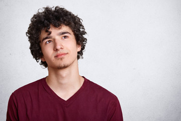 Portrait of thinking young man with dark curly hair, stands with thoughtful facial expression, dresses maroon shirt. Student dreams isolated over white concrete wall. Youth and teenagers concept.