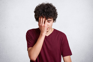 Disappointed man in maroon t shirt covers his face with one hand over white background. Handsome...