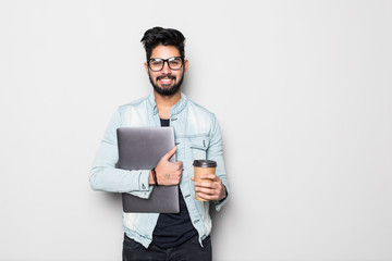 Portrait of handsome casual Indian man drinking coffee while holding laptop computer, standing on white background