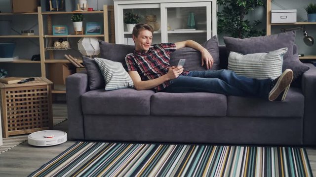 Smiling guy is sufring the Internet with smartphone relaxing on sofa while robotic vacuum cleaner is cleaning floor removing dust from carpet. Gadgets and housekeeping concept.
