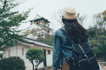 Fototapeta na wymiar Back view of young asian woman traveler wear hat with backpack on shoulder out sightseeing foreign old city in japan. female chinese examines architectural monument osaka castle standing in garden.