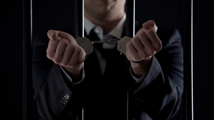 Man in suit showing hands in handcuffs behind bars, bribery, financial fraud