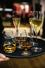 Young elegant man standing in the restaurant, holding a plate with glasses of wine and cognac, whisky. Man's style