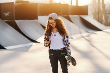 Skinny mixed race teenage urban girl using smart phone and holding skateboard while standing at skate park.