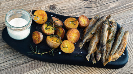 fried breaded capelin, halved young potatos, and rosemary on a wooden serving board with a jar of tartar sauce on side placed on a rustic table top