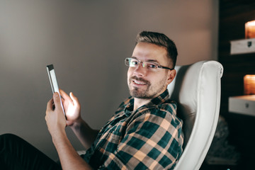 Portrait of smiling handsome Caucasian unshaven employee using tablet while sitting in office late at night.