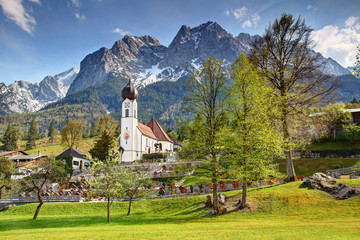 Small Bavarian domed church St Johannes, graveyard, houses and blue sky in spring with Waxenstein peak Wetterstein range Northern Limestone Alps Grainau Garmisch Partenkirchen Bayern Germany Europe
