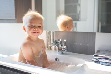 Baby taking bath in sink. Child playing with foam and soap bubbles in sunny bathroom with window....
