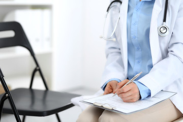 Female doctor writing up medication history records form on clipboard, while sitting at the chair. Physician at work in hospital or clinic. Healthcare, insurance and medicine concept