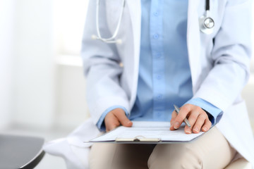 Female doctor writing up medication history records form on clipboard, while sitting at the chair. Physician at work in hospital or clinic. Healthcare, insurance and medicine concept