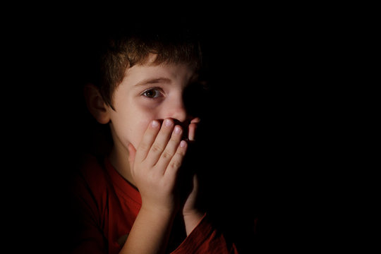 Portrait in the dark of a young scared sad boy of 7 years scared and upset hands on the mouth in the dark backround in the profile.