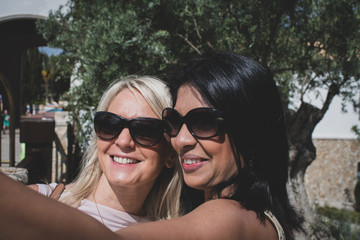 An interracial lesbian couple taking a selfie and laughing whilst looking at the photograph on a bright sunny day outdoors. 