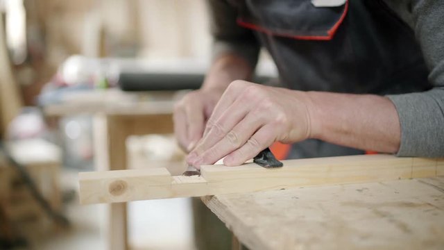 Closeup hands of carpenter attaching wooden plank to workbench with clamps and cutting off small piece of wood with chisel