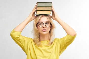 Smart beautiful young blonde in glasses holds books over her head with a serious face in yellow clothes alone in a white studio background