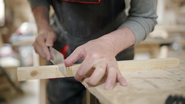 Hands of carpenter sawing wooden plank attached to workbench with clamps and using chisel to cut off small pieces of wood