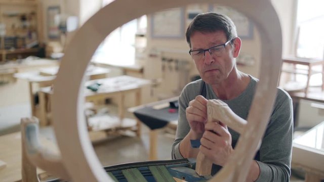 Medium shot of experienced mature furniture maker in eyeglasses using sandpaper for sanding arm of handmade vintage chair in his studio, through object view