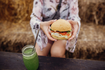 Stylish hipster girl holding delicious vegan burger and smoothie in glass jar in hands at street food festival, close up. Happy boho woman tasting burger with drink in summer street. Zero waste