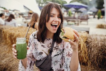 Stylish hipster girl in sunglasses holding delicious vegan burger and smoothie in glass jar in...