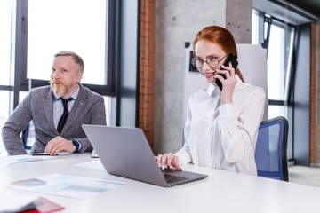 Red-haired girl with glasses office worker talking on the phone sitting at a table in the office on the background of an adult man with gray hair