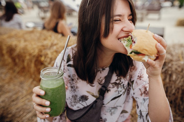 Stylish hipster girl in sunglasses eating delicious vegan burger and holding smoothie in glass jar in hands at street food festival. Happy boho woman biting burger with drink in summer street