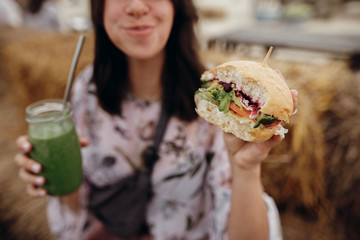 Stylish hipster girl holding delicious vegan burger and smoothie in glass jar in hands at street...