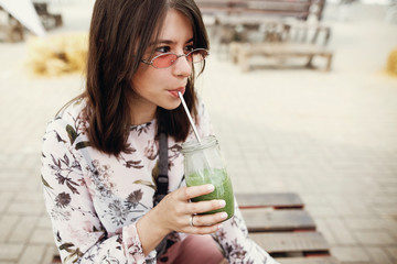 Stylish hipster boho girl drinking spinach smoothie in glass jar with metal reusable straw at street food festival. Happy woman in sunglasses with healthy drink in summer street. Zero waste