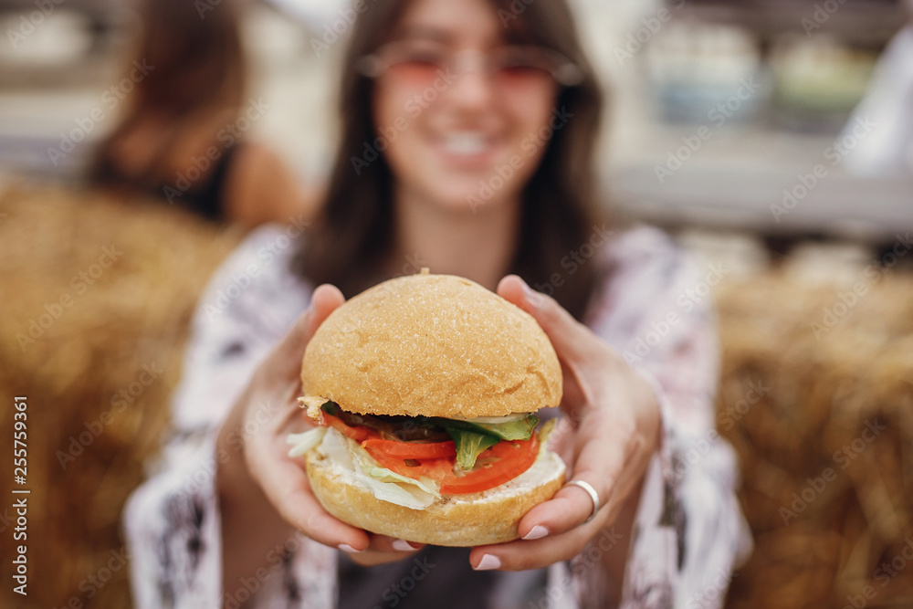 Wall mural Vegan burger closeup in boho girl hands. Stylish hipster girl in sunglasses eating delicious vegan burger at street food festival. Happy boho woman tasting and biting burger