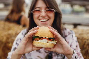 Stylish hipster girl in sunglasses eating delicious vegan burger at street food festival. Happy boho woman tasting and biting burger with vegetables in summer street. Street food festival