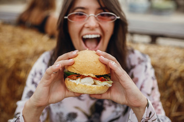 Street food festival. Stylish hipster girl in sunglasses eating delicious vegan burger at street food festival. Happy boho woman tasting and biting burger with vegetables in summer street.