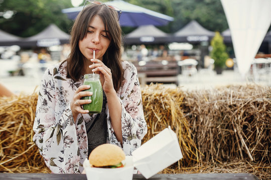 Stylish Hipster Boho Girl Drinking Smoothie In Glass Jar With Delicious Vegan Burger On Wooden Table At Street Food Festival. Happy Woman With Healthy Drink In Summer Street. Zero Waste