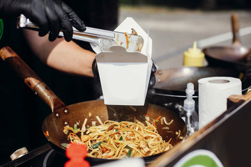 Fried chinese japanese noodles with vegetables and shrimps in takeaway box.Food delivery. Chef putting noodles in carton box to go from open kitchen. Asian street food festival