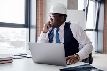African Builder in a white helmet sitting at a table in the office. Big boss of the construction...
