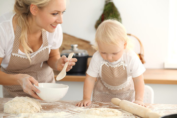Little girl and her blonde mom in beige aprons  playing and laughing while kneading the dough in kitchen. Homemade pastry for bread, pizza or bake cookies. Family fun and cooking concept