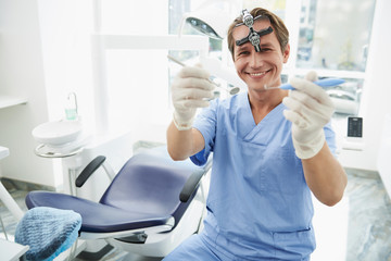 Cheerful dentist demonstrating instruments while sitting near dental chair