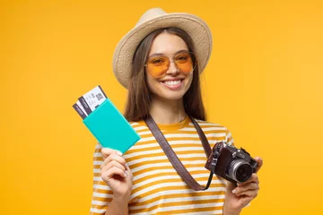 Deurstickers Excited cheerful young tourist girl holding passport with tickets and camera, isolated on yellow background © Damir Khabirov